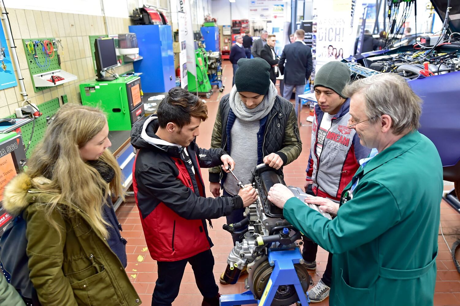 Vier Jugendliche stehen in der KFZ-Werkstatt. Ein Ausbilder der HWK Chemnitz zeigt ihnen einen Motorblock. Im Hintergund ist ein Fahrzeug zu sehen und noch andere Teilnehmer am Tag der Bildung, die die Werkstätten besichtigen.