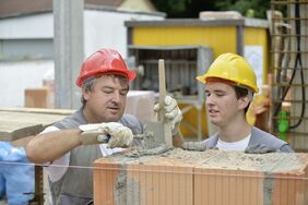 Ein Ausbilder im Maurergewerk zeigt einem Maurerlehrling, wie eine Mauer fachgerecht gesetzt wird.
