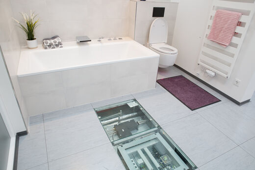 Bad mit Badewanne, Toilette und Heizungshandkörper im E-Haus. Zwei Bodenfliesen sind durchsichtig, so dass die verlegten Leitungen sichtbar sind.