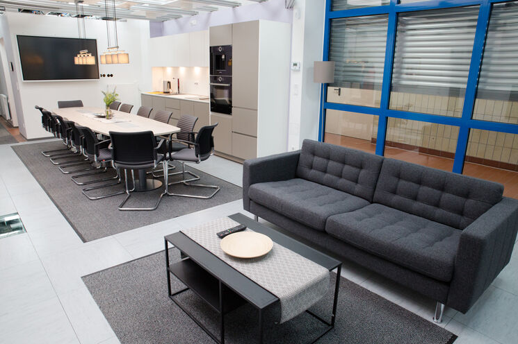 Im Vordergrund eine eine graue Couch mit einem grauen Wohnzimmertsch zu sehen. Im Hintergrund ist die offene Küchenzeile und ein Besprechungstisch mit 12 Stühlen zu sehen. Außerdem hängt an der Stirnseite des Besprechungstisches ein Fernseher an der Wand.