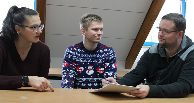 Drei Personen sitzen am Tisch. Einer erklärt etwas mit Zeigen auf Block.