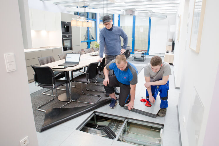 Ausbildungssituation. Zwei Lehrlinge knieen auf dem Boden und schauen sich die verlegten Rohre und elektrischen leitungen an. Der Ausbilder steht hinter den Lehrlingen. Im Hintergrund sieht man einen Teil der Küche und des Besprechungstisches.