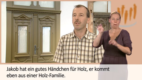Auf dem Bild ist im Hintergrund eine Hauseingangstür zu sehen. In der Mitte des Bildes steht ein Mann im karierten T-Shirt und erklärt, warum es toll ist in diesem Unternehmen zu arbeiten. Am Rand des Bildes ist eine Frau zu sehen. Diese spricht die Gebärdensprache.