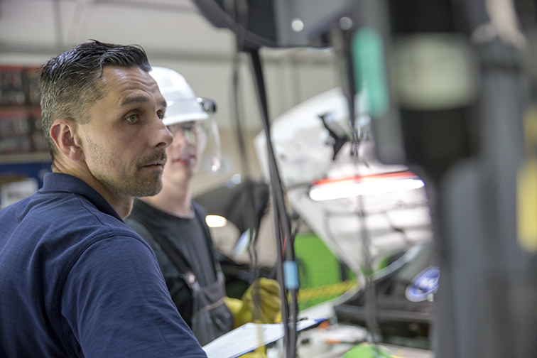 Das Bild zeigt eine Ausbildungssituation in einer Werkstatt. Zwei Männer stehen an einer Maschine und beobachten einen Prozessablauf.