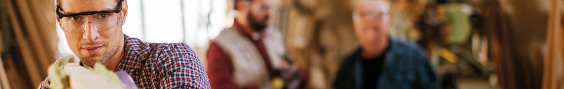 Auf dem Bild ist ein Mann mit Schutzbrille und Arbeitsschutzhandschuhen in einer Werkstatt abgebildet, der ein Stück Holz in der Hand hält und dieses hinsichtlich seiner Beschaffenheit prüft. Im Hintergrund sind verschwommen zwei weitere Männer im Gespräch erkennbar.